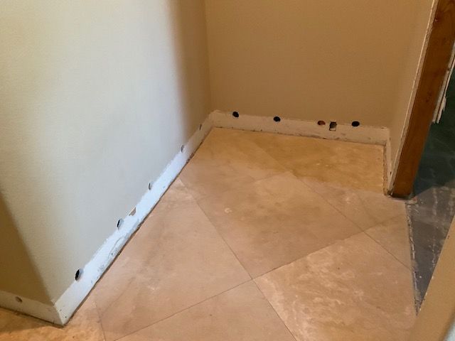 Corner of a room with beige tile floor, tan walls, and a doorway. Black holes line the baseboards.