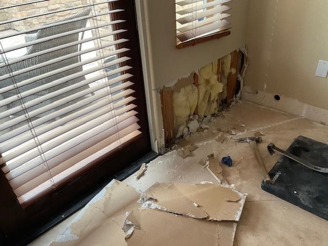 Wall damage near a window, revealing insulation and wood framing. Debris and tools litter the floor.