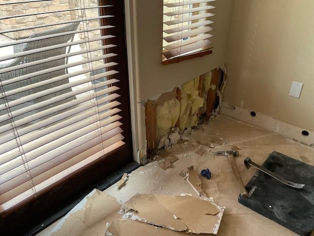 Wall damage near a window. Drywall is ripped away, revealing insulation and wood framing. Debris on floor.