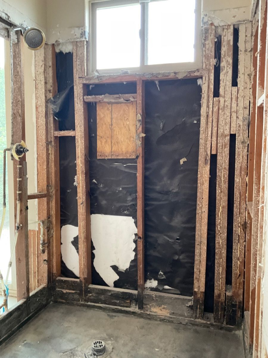 Bathroom shower wall under construction, revealing studs, black moisture barrier, and window.