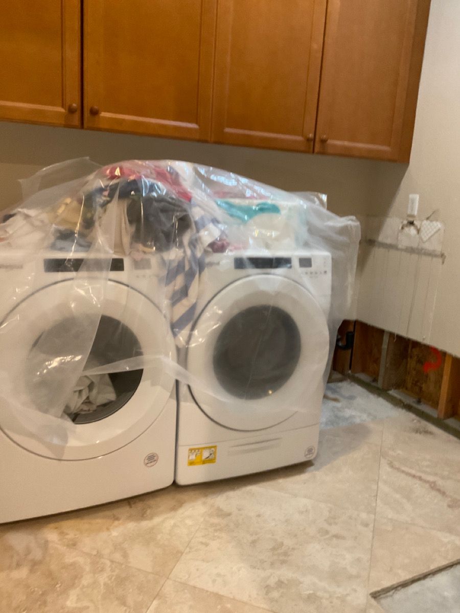 White washer and dryer under plastic sheeting in a room with wood cabinets and partially exposed wall.