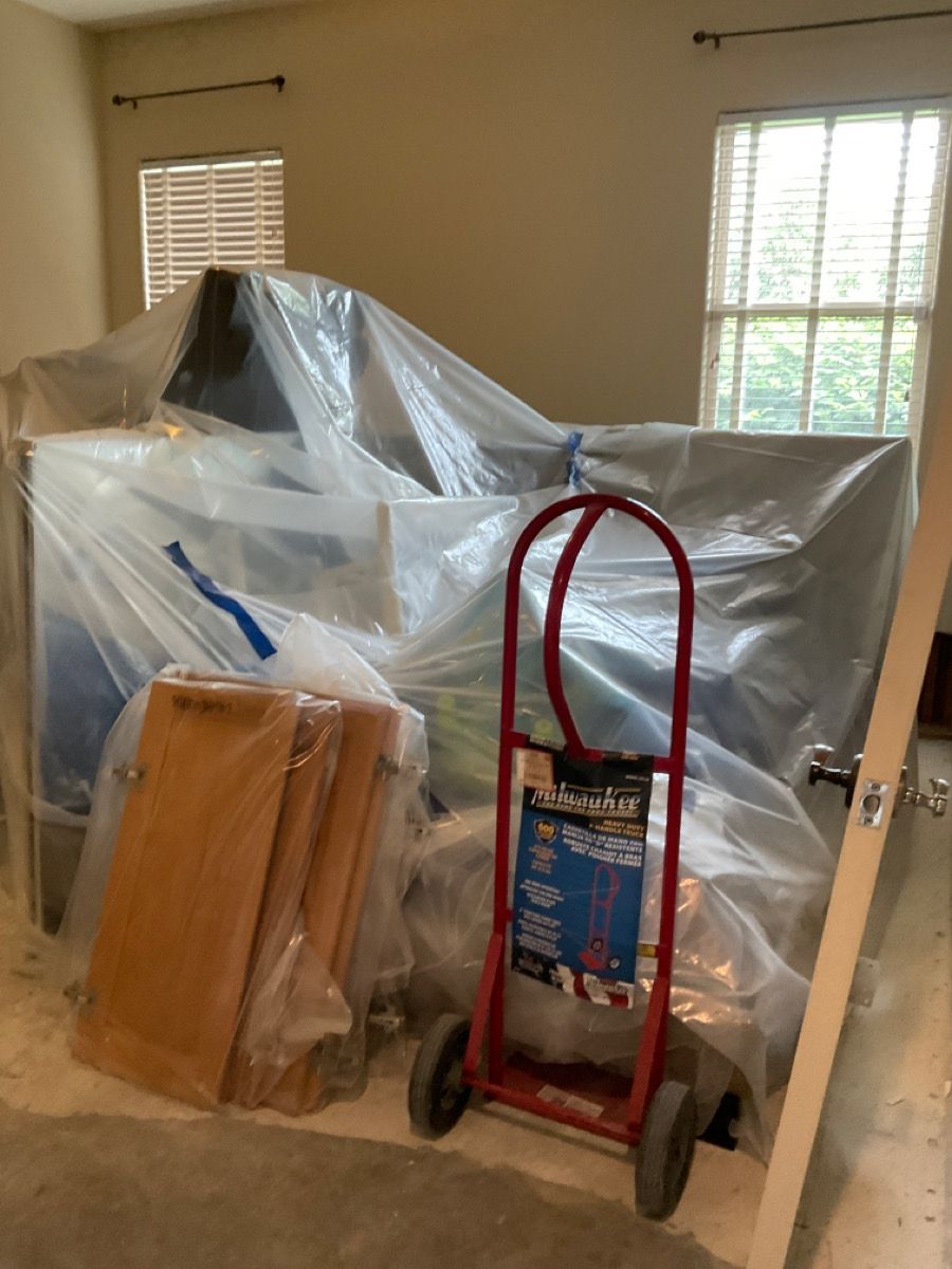 Room with furniture covered in plastic, red hand truck, wooden cabinet doors, and windows.