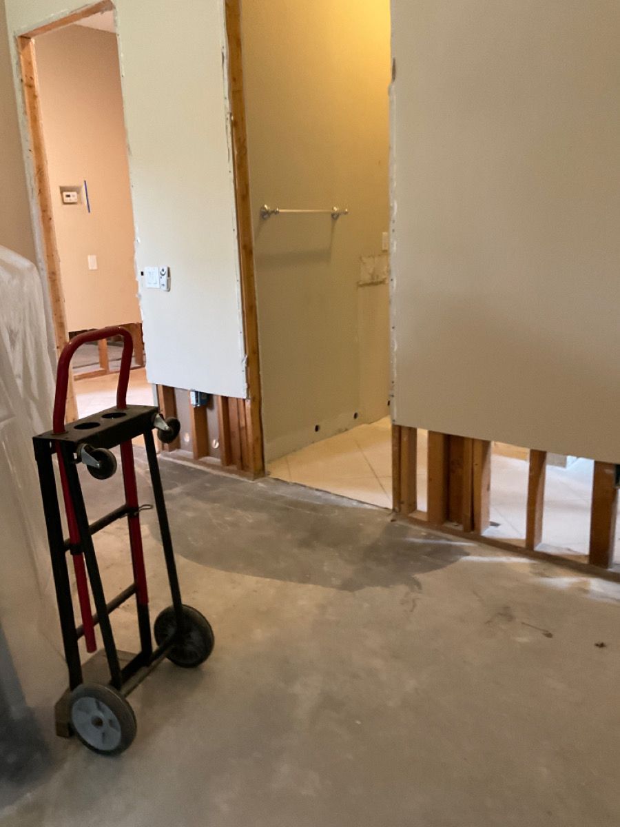 A room under construction with exposed studs, drywall, and a hand truck; a bathroom doorway is visible.