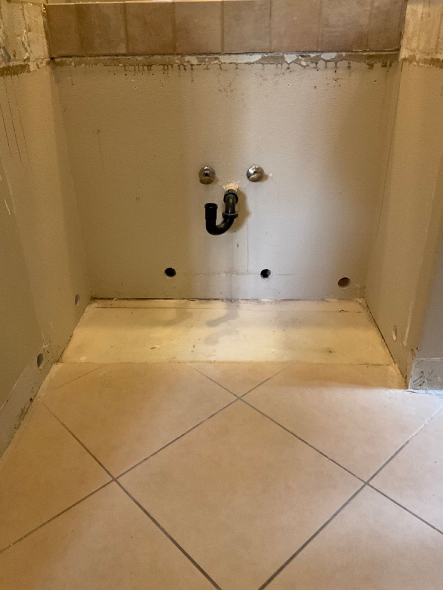 Bathroom sink wall with exposed plumbing. Beige tile floor and wall, two chrome valves, black drain pipe.