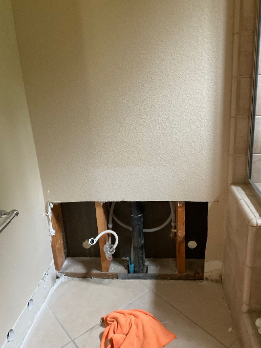 Hole in bathroom wall reveals plumbing, pipes, and studs. Orange towel on tiled floor.