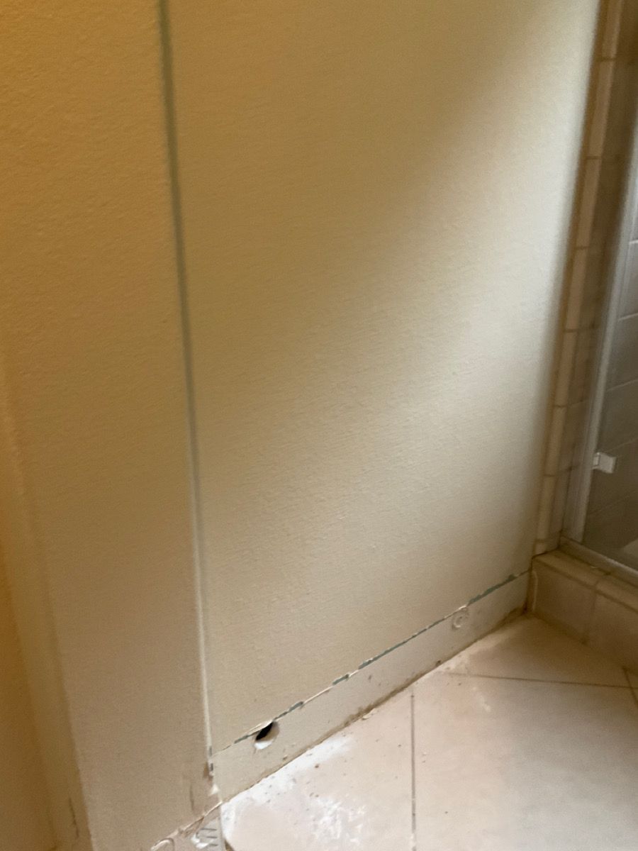 Beige wall with a small hole near the base, alongside a tiled floor and shower door.