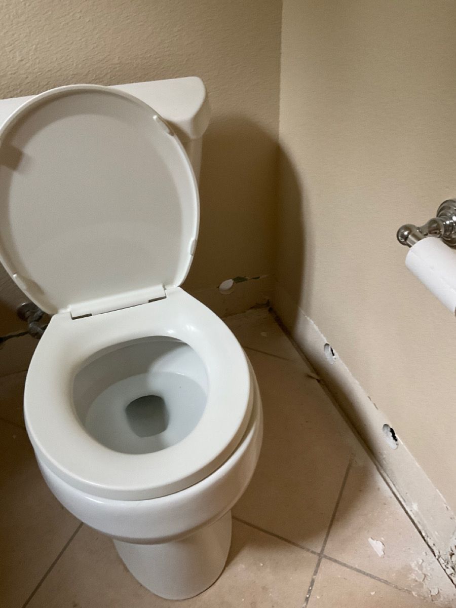 Toilet with the lid up in a beige bathroom. Damaged wall next to the toilet, toilet paper dispenser visible.