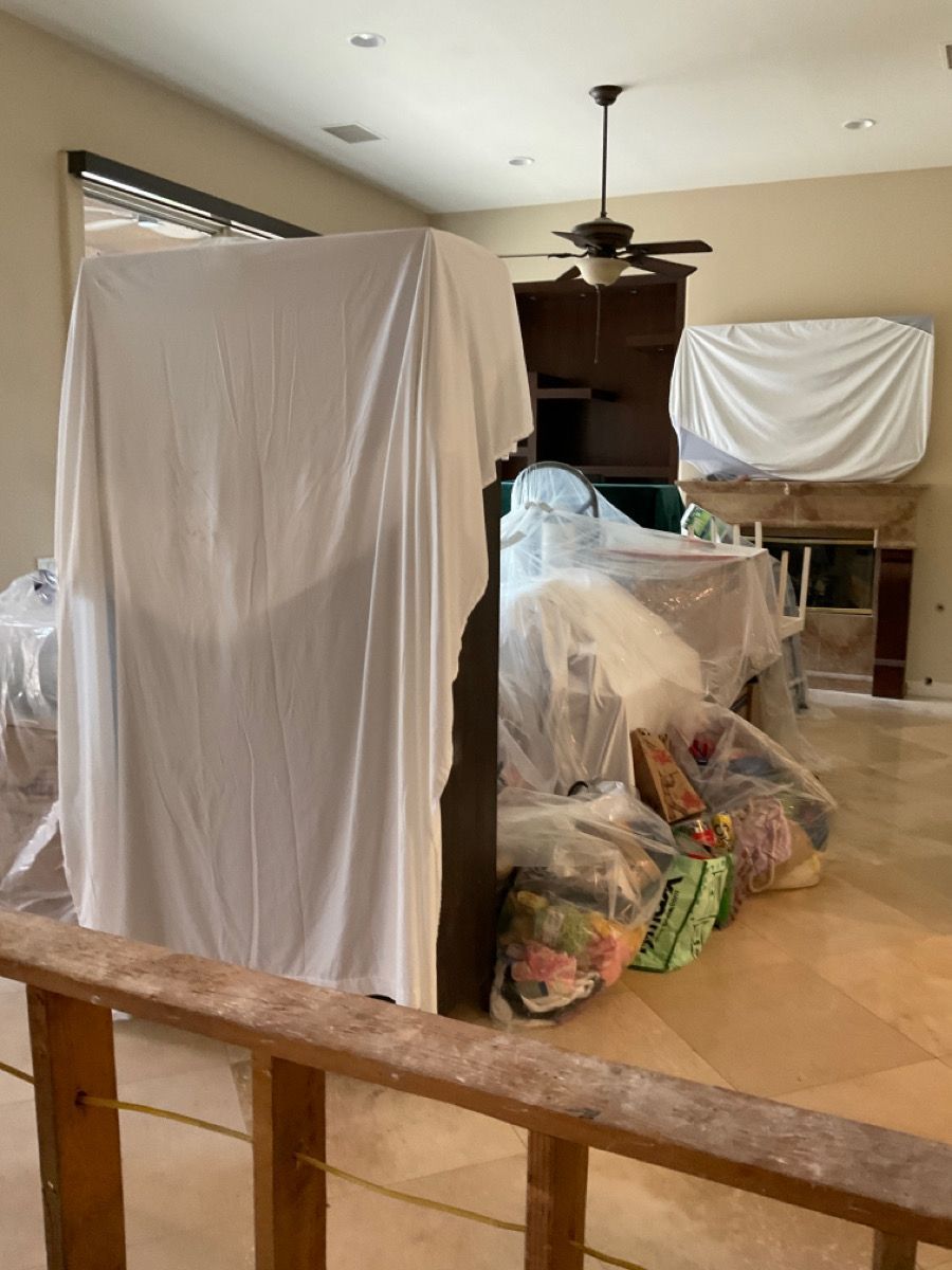 Room with furniture covered in white sheets. Bags and items piled on floor. Ceiling fan visible.