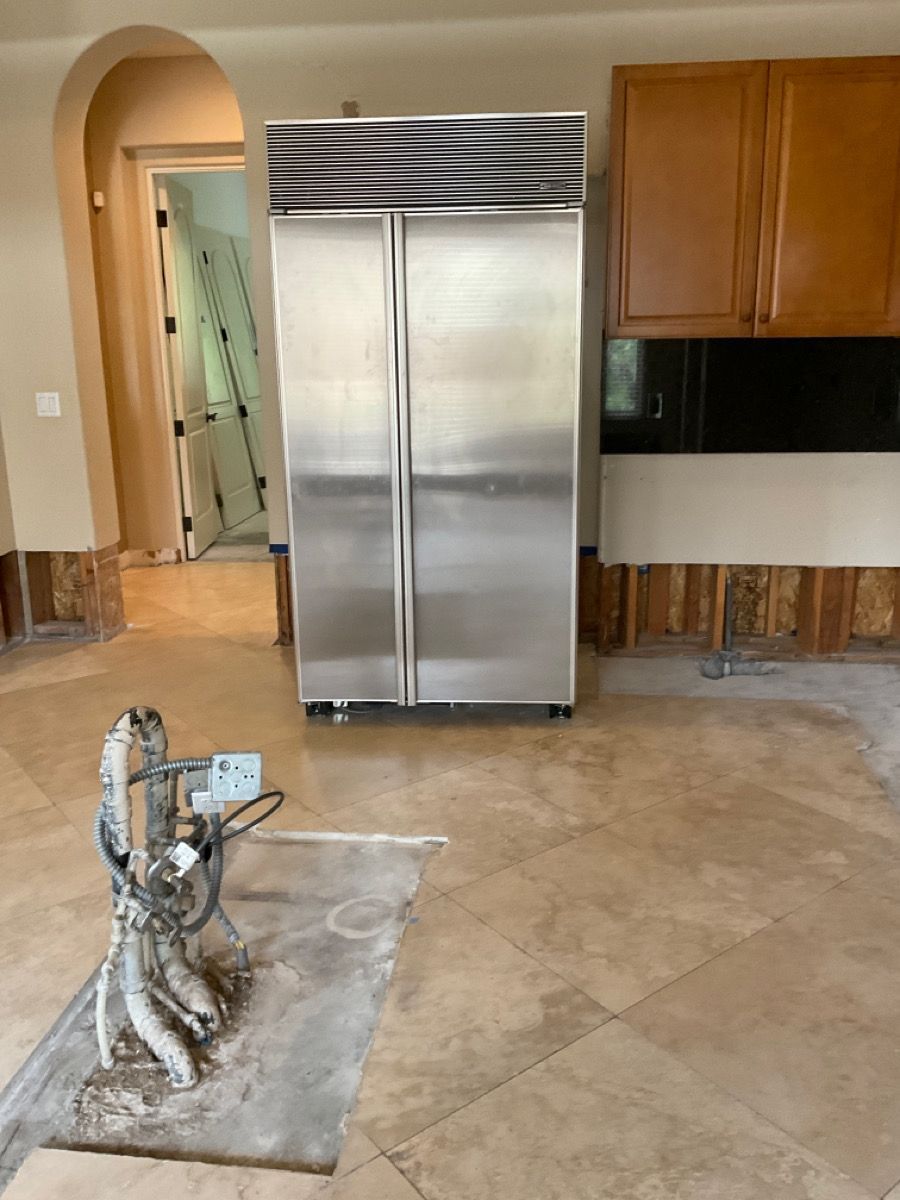 A stainless steel refrigerator stands in a kitchen under renovation. A tile cutter is on the floor.