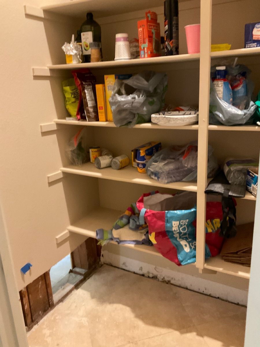 Pantry with various food items on shelves. Lower shelf has clutter, exposed floor damage.