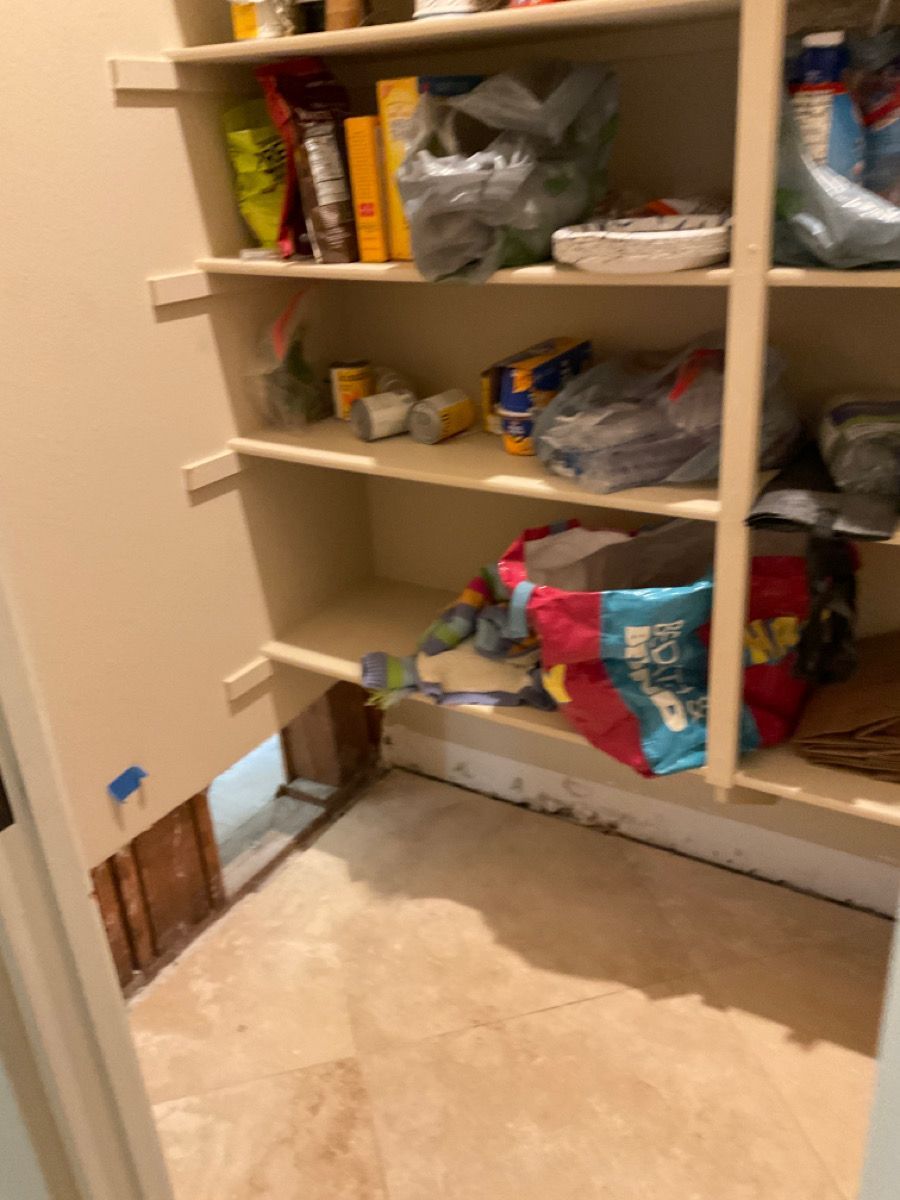 Pantry interior with exposed foundation, shelving stocked with food items, and a door open.