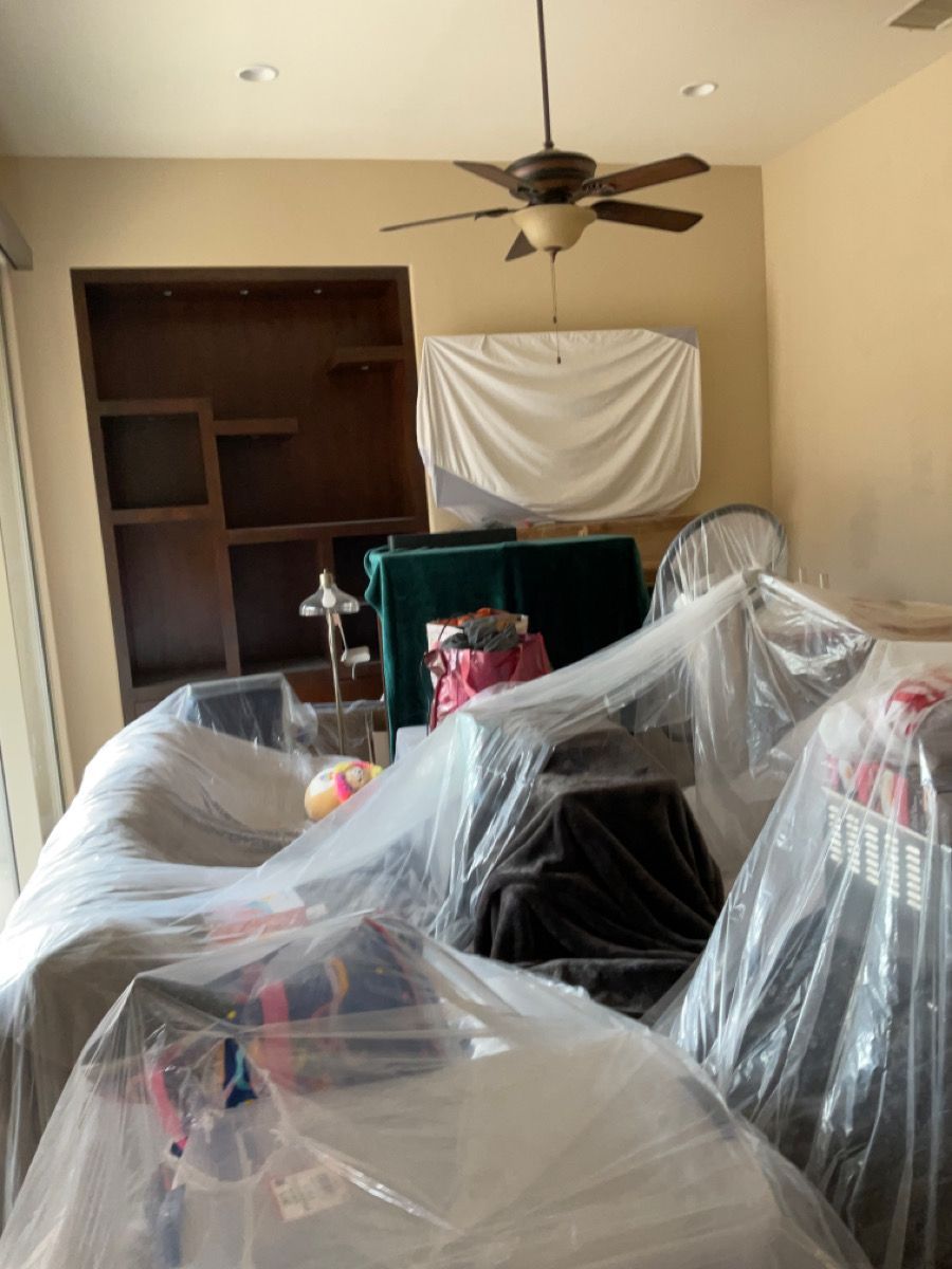 Living room filled with furniture covered in plastic wrap, ready for moving. Brown built-in bookshelf, beige walls.