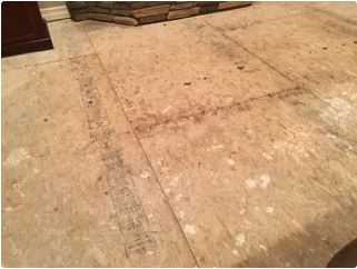 Bare, stained plywood floor with dark markings, near a fireplace and wall trim.