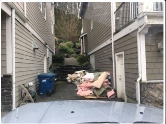 Narrow alley between two houses, filled with construction debris and a blue trash bin.