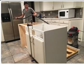 Man vacuums a white kitchen cabinet in a kitchen with stainless steel refrigerator and microwave.