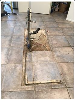 Hole cut in tiled floor, revealing plumbing. Metal pipes and wires protrude.