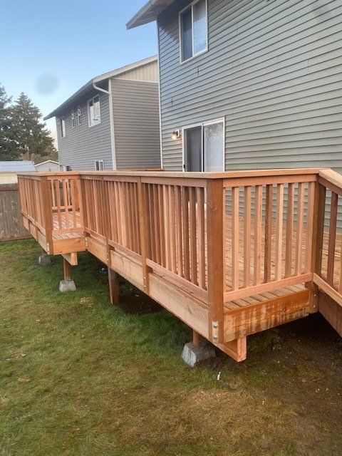New wooden deck attached to a two-story house, with green grass and a gray building in the background.