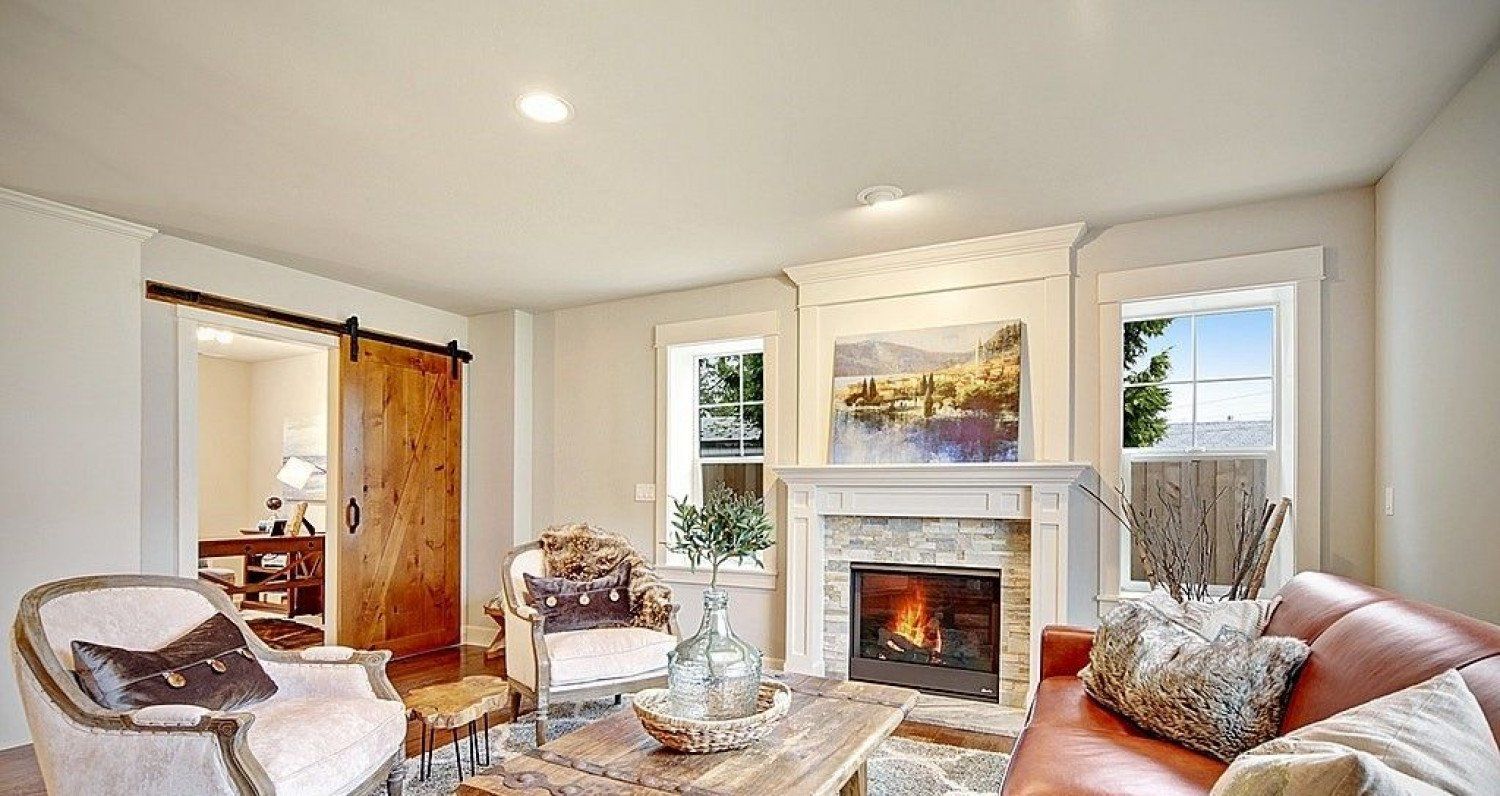 Cozy living room with fireplace, leather sofa, wooden barn door, two windows, and two patterned armchairs.