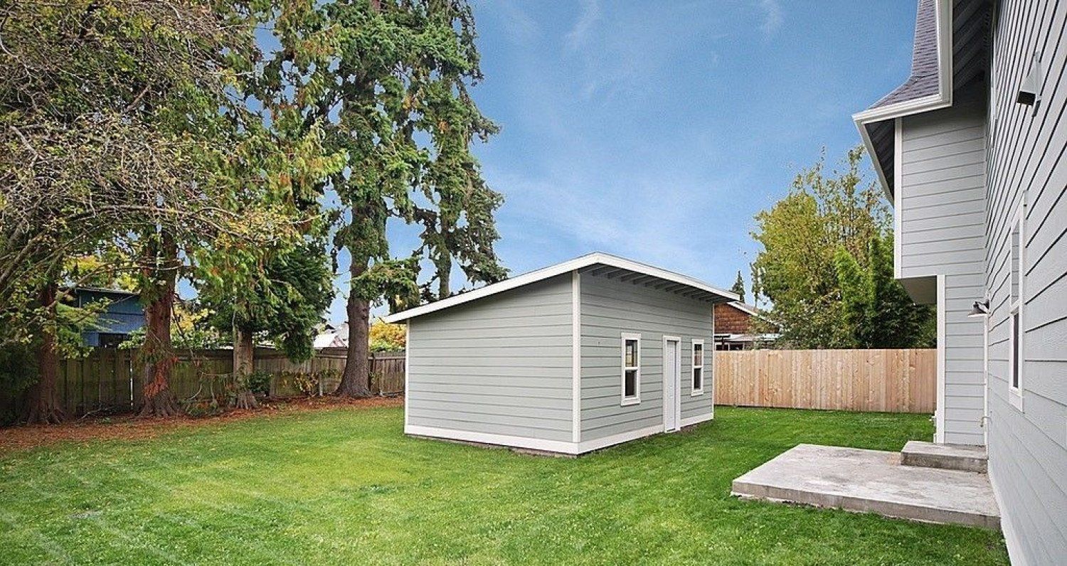 Gray shed in a grassy backyard, trees and a house on a sunny day.