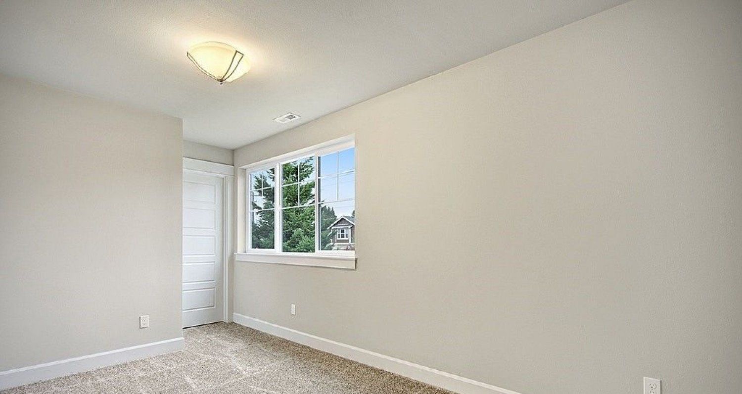 Empty bedroom with a window, door, and light-colored walls and carpet.