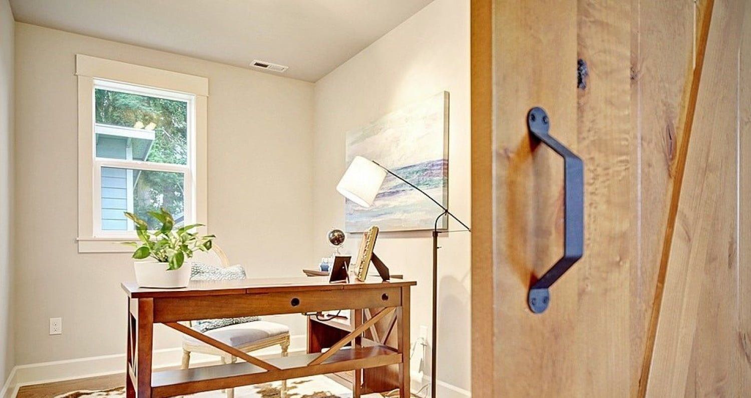 Wooden desk in a home office near a window and open door. Beige walls, wood door with black handle.