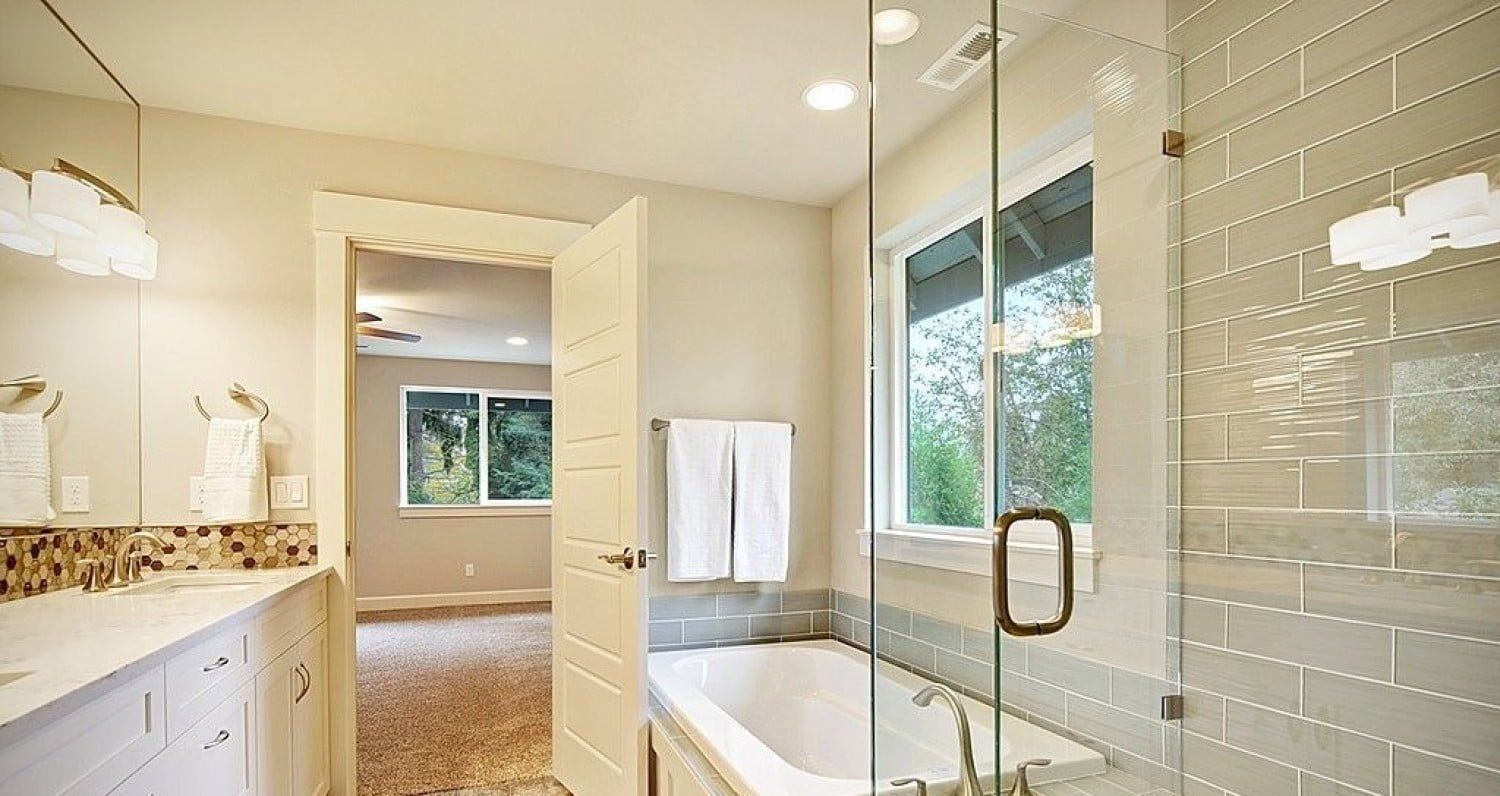 Bathroom with glass shower, soaking tub, window, and door to bedroom with neutral colors and modern design.