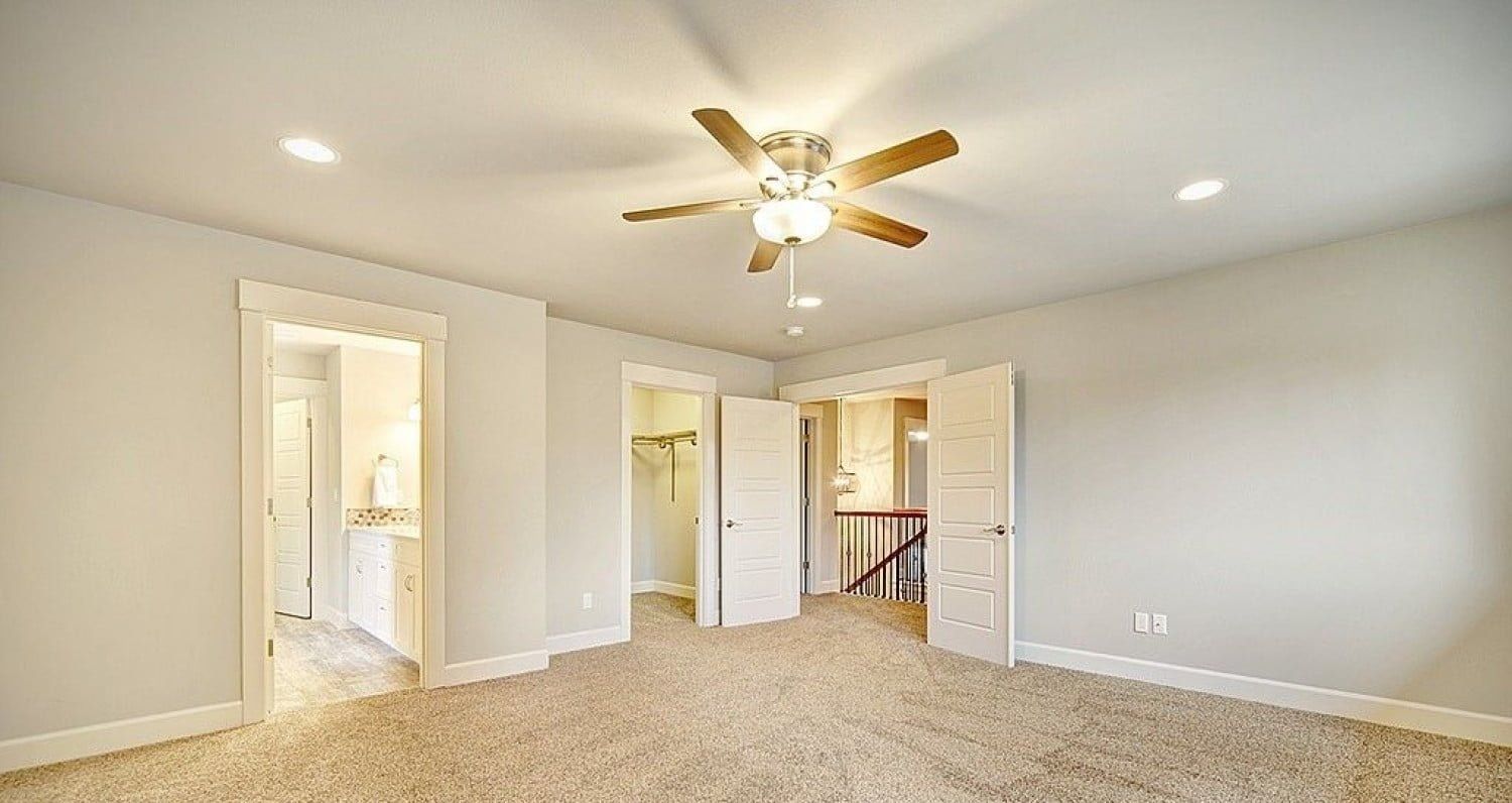 Empty bedroom with light beige walls, tan carpet, and a ceiling fan. Three doorways lead to other rooms.