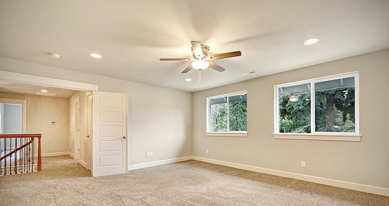 Empty bedroom with beige walls, carpet, two windows, and a ceiling fan.