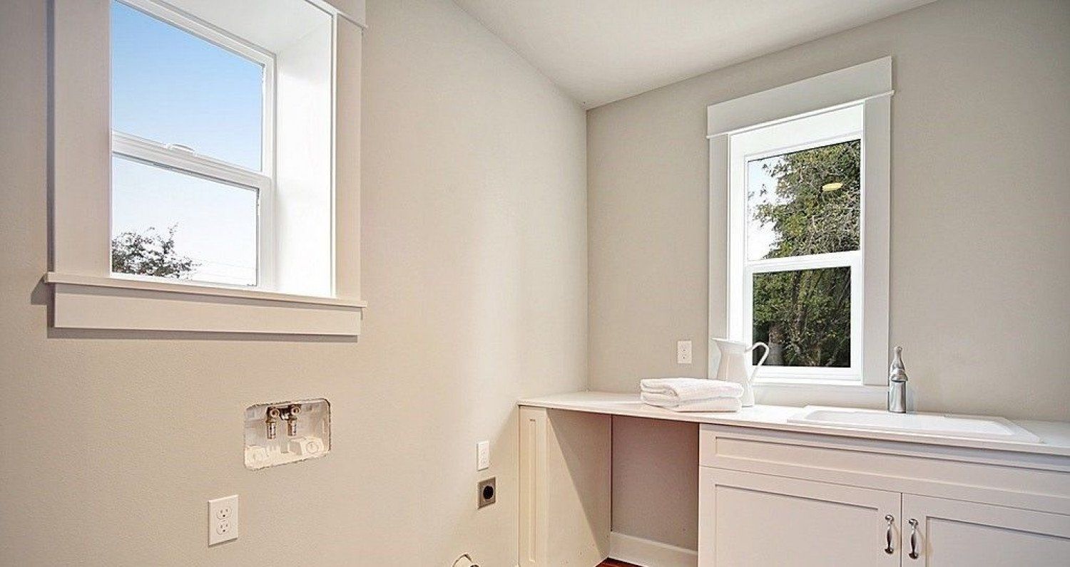 Empty laundry room with white cabinets and trim, two windows, and electrical outlets.