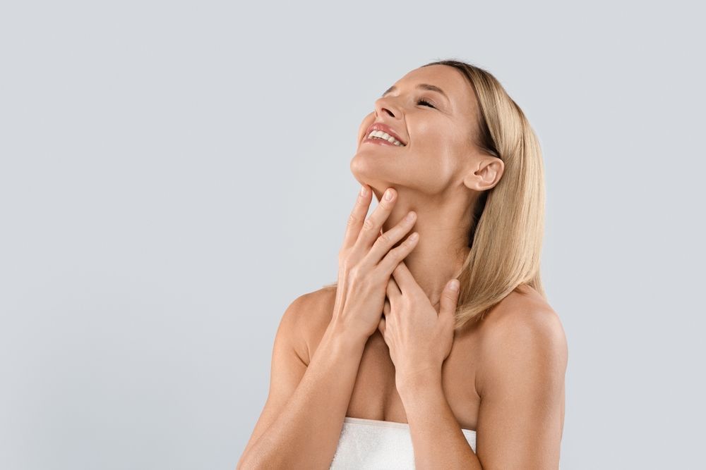 Woman in towel smiles, touching her neck, eyes closed, against a light gray background.