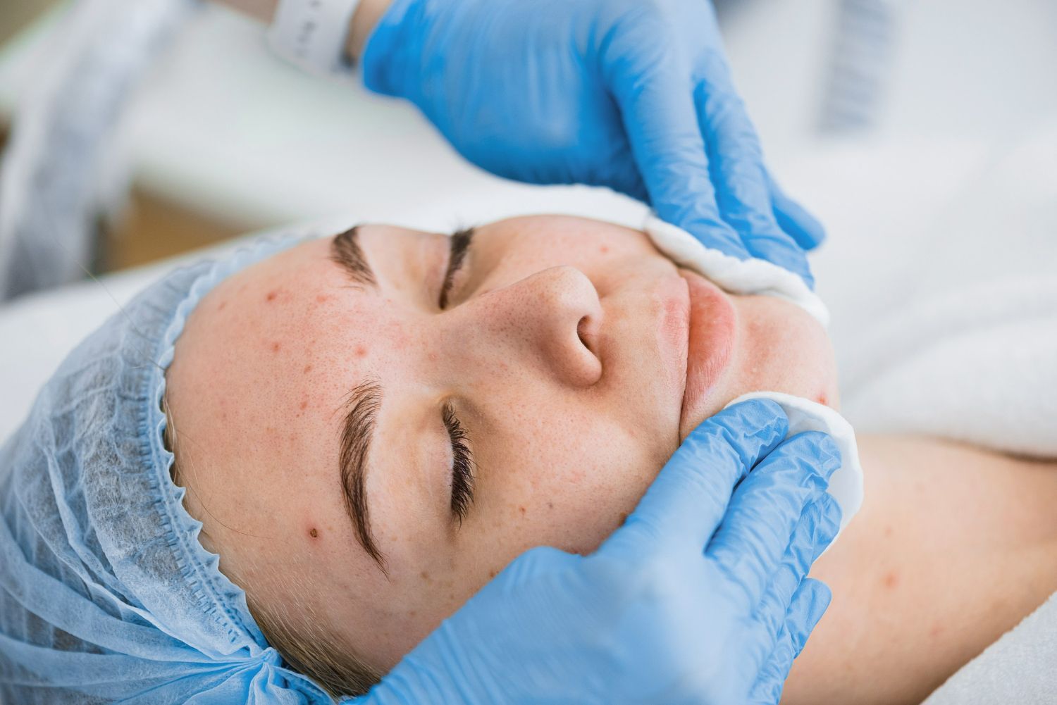 A professional in a white coat examines a client’s skin as the client views their reflection in a hand mirror.