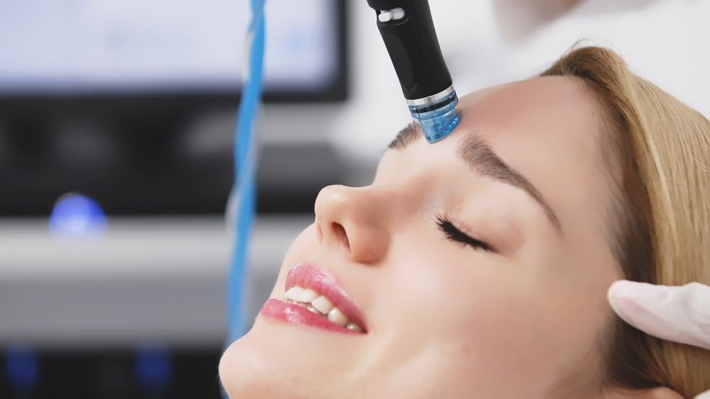 Woman receiving a facial treatment with eyes closed, a device held near her forehead. The setting appears to be a spa.