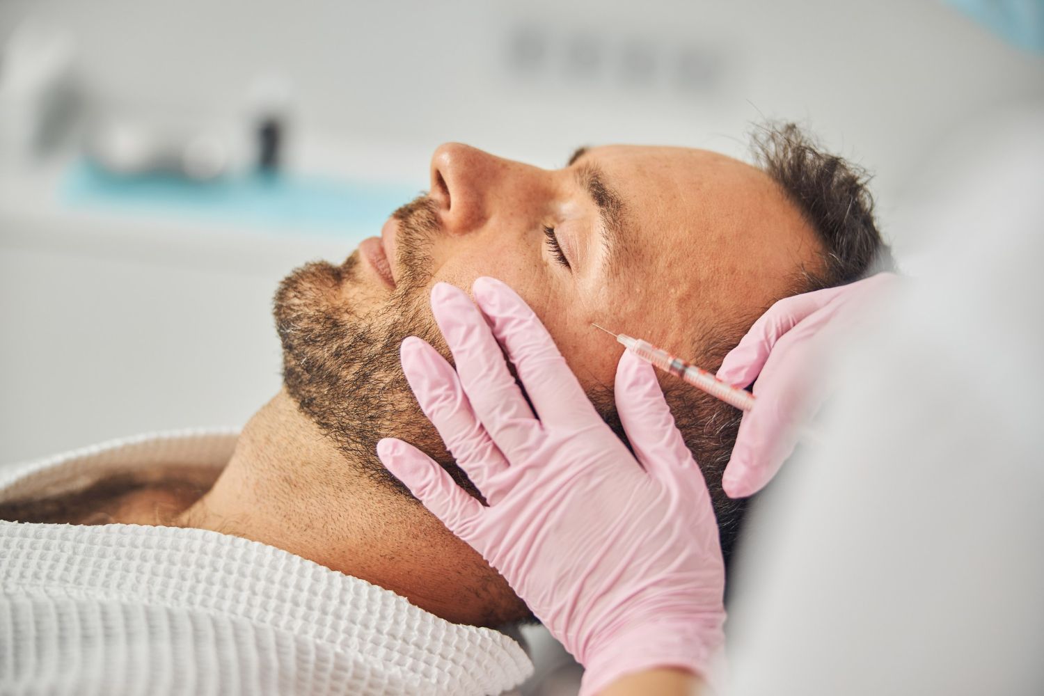 A person in a robe receives a cosmetic facial injection from a practitioner wearing pink medical gloves.
