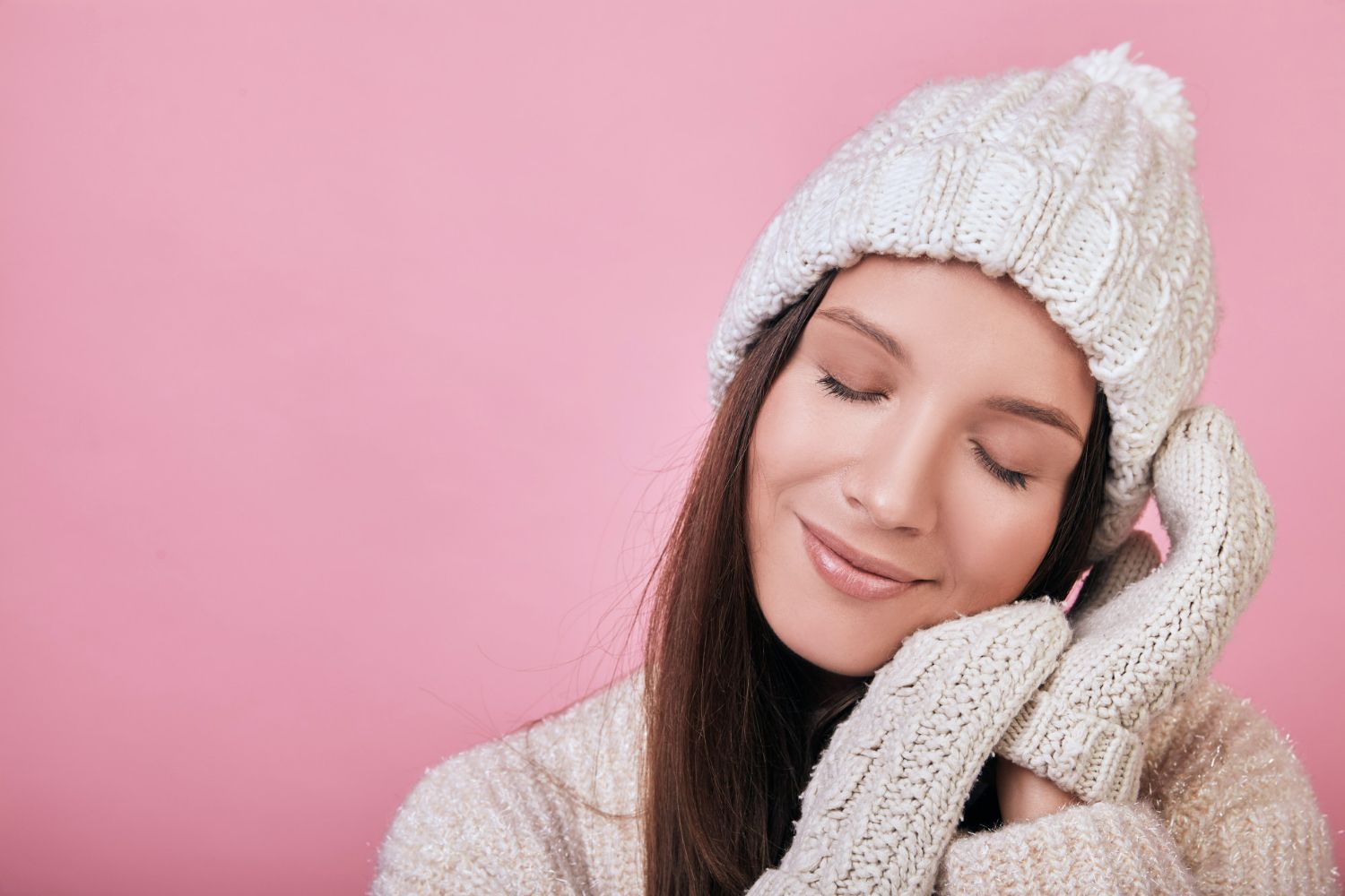 Woman in white knit hat, gloves, and sweater smiles with eyes closed, pink background.