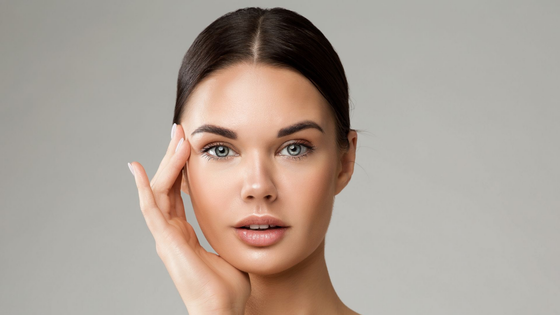 Woman with fair skin, dark hair pulled back, touching her face, against a gray background.