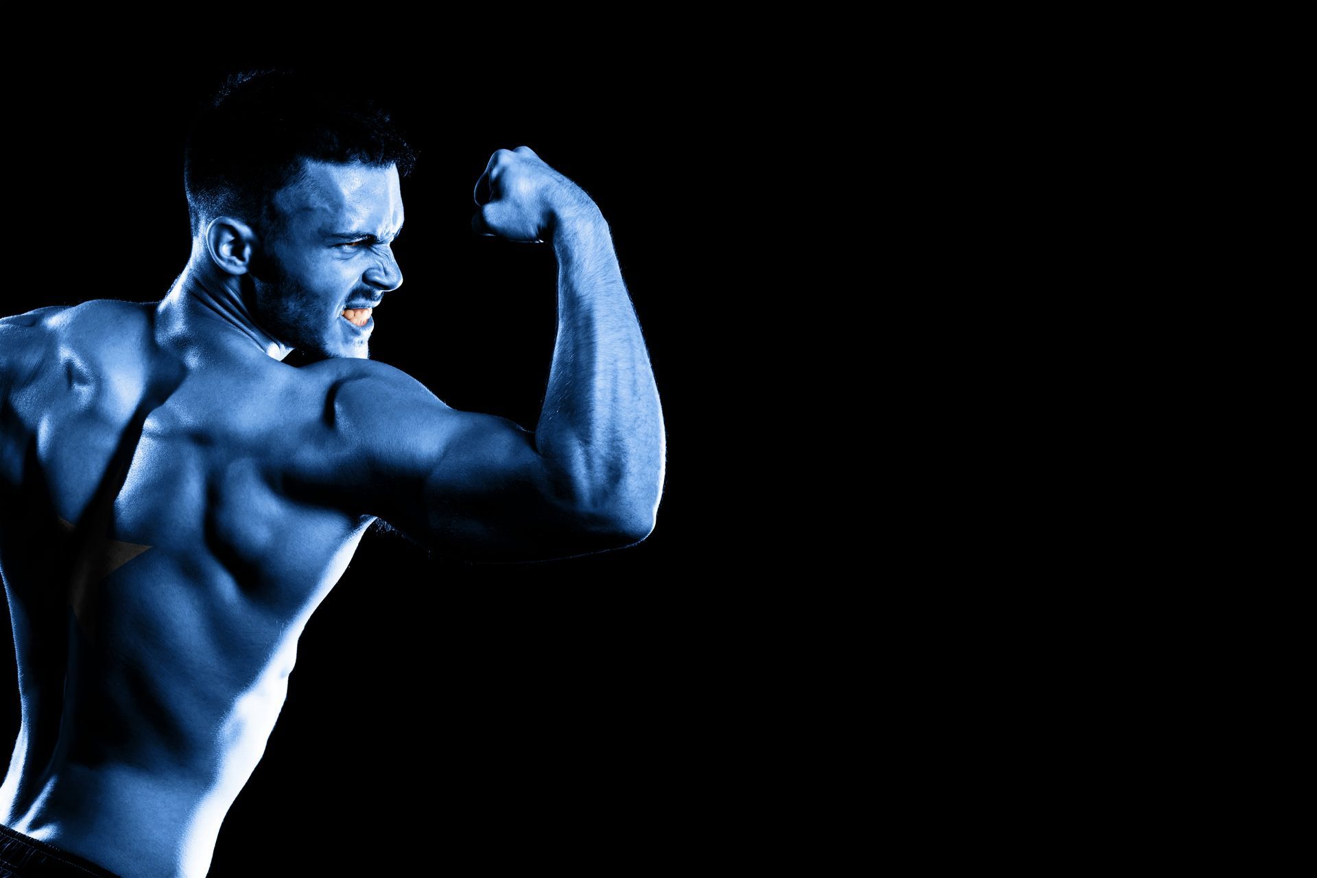 A shirtless man is flexing his muscles in a dark room.