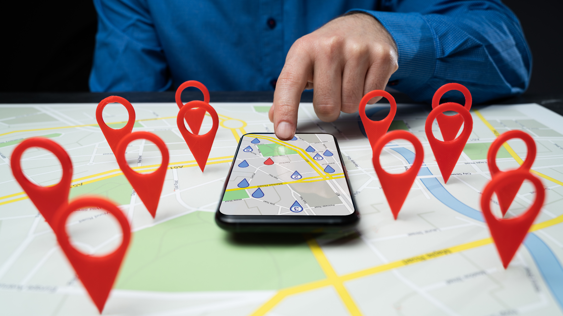 Smartphone on a map surrounded by red location pins, with a person pointing nearby.