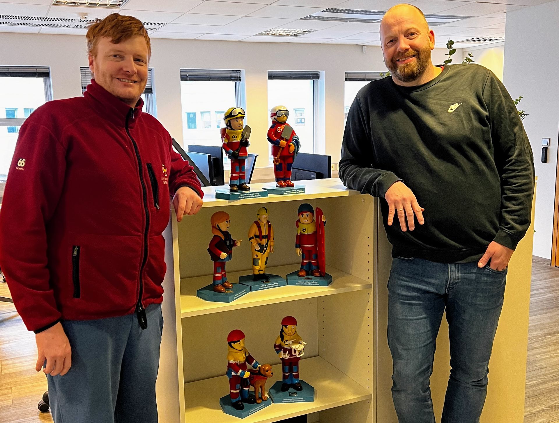 Two men are standing in front of a shelf with figurines on it.