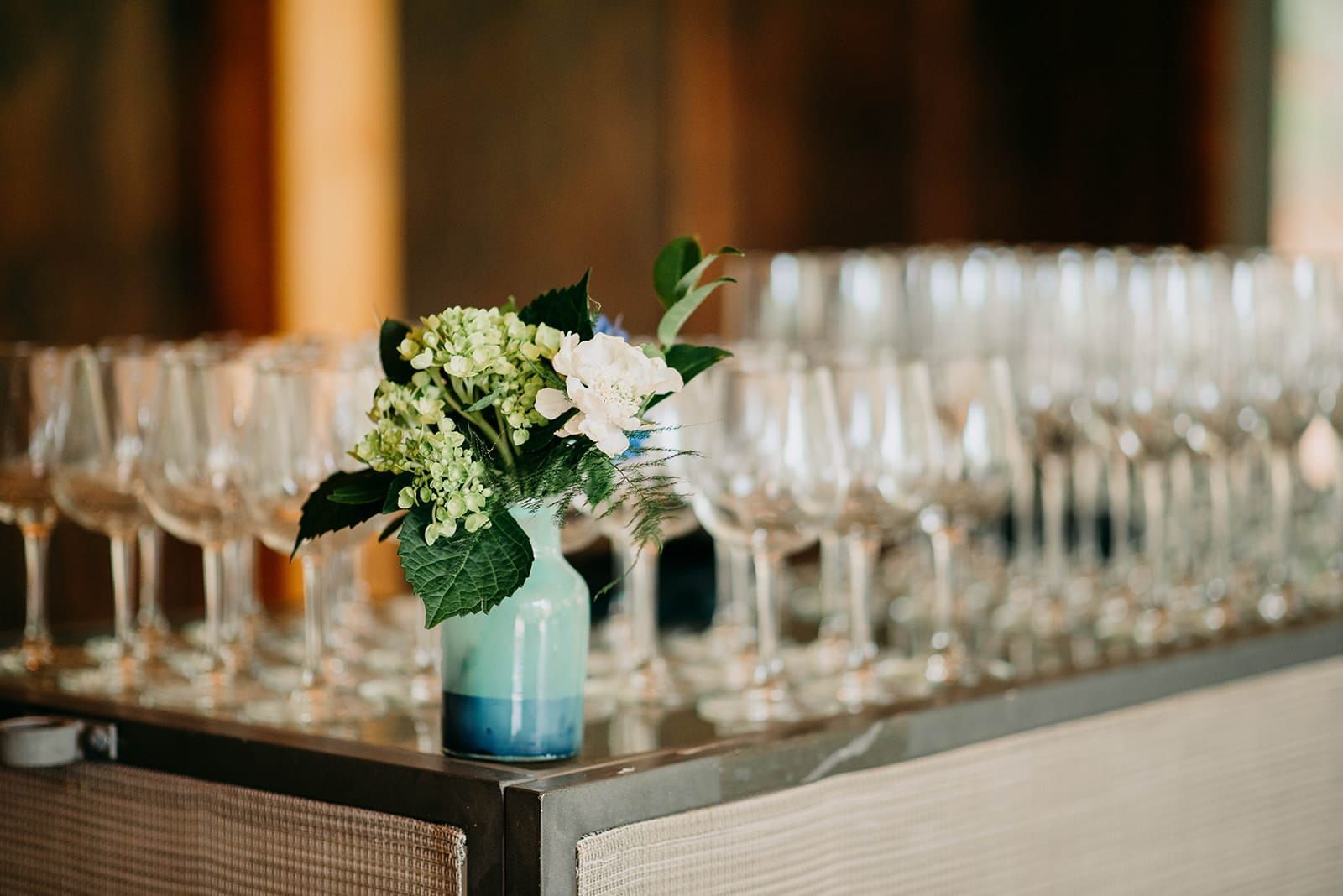 A small vase of flowers sits on a bar, in front of a row of wine glasses.
