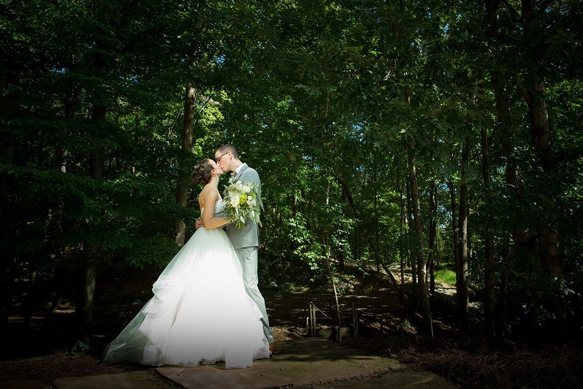 Bride and groom kissing in a forest setting, the bride in a white gown, the groom in a gray suit.