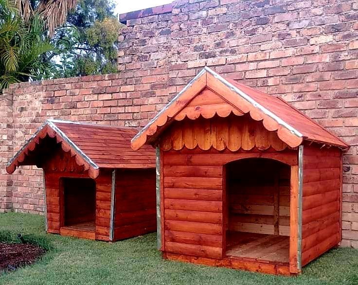 Two wooden dog houses are sitting in front of a brick wall and are available from Pets and the City in Pretoria East.