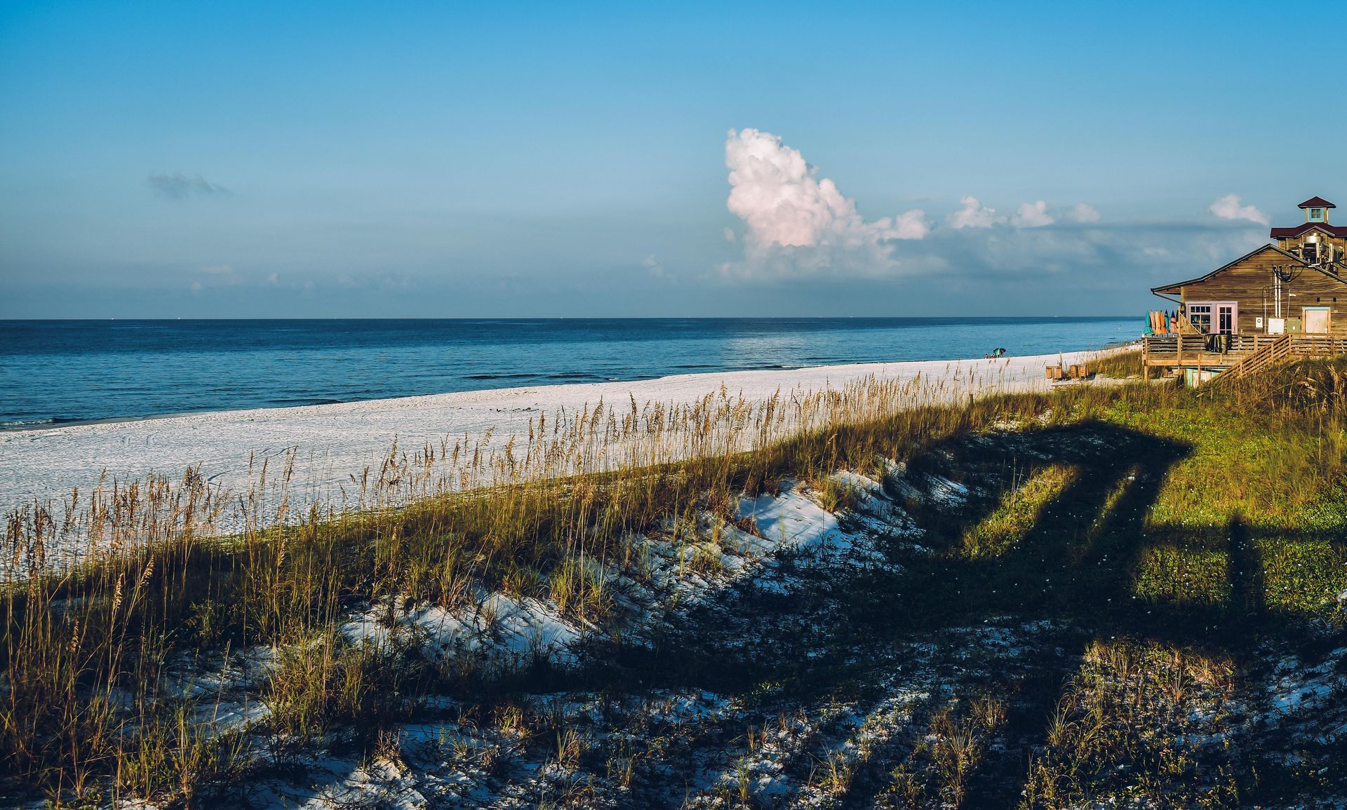Beach scene with white sand, blue water, and a weathered wooden building under a partly cloudy sky.