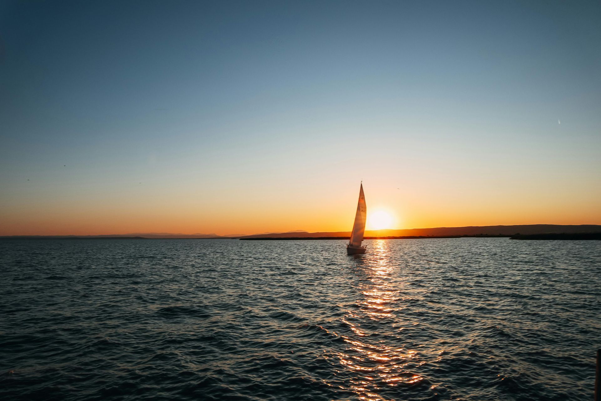 Sailboat on water at sunset, silhouetted against orange and blue sky.