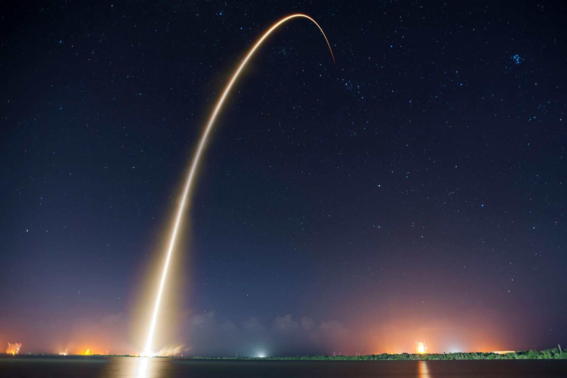 SpaceX Rocket Launch, Brevard County FL. Rocket launches into the night sky, leaving a bright trail. Coastal area lit up below, with stars overhead.