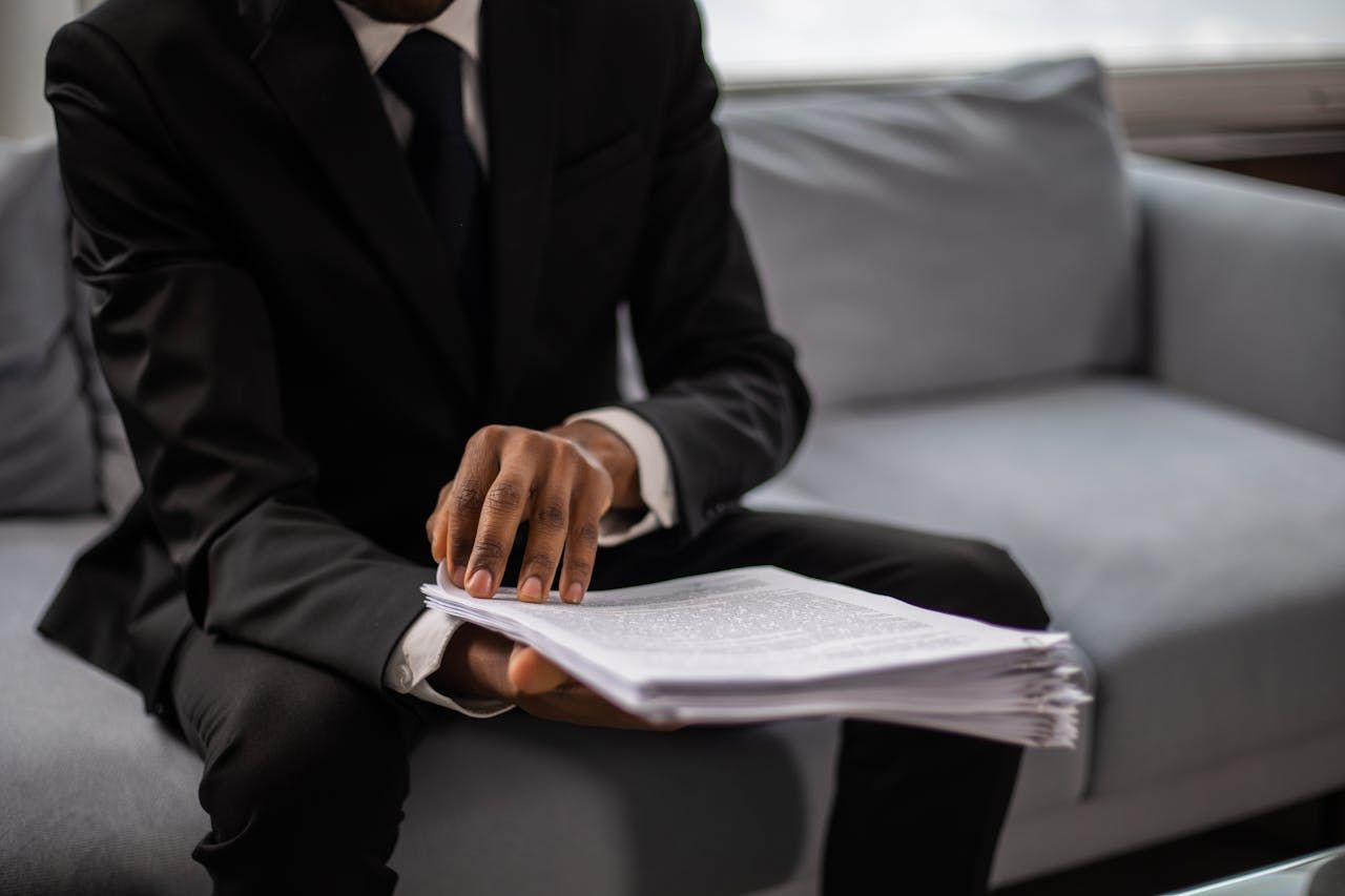 Person in black suit holding a stack of papers while seated on a gray couch.