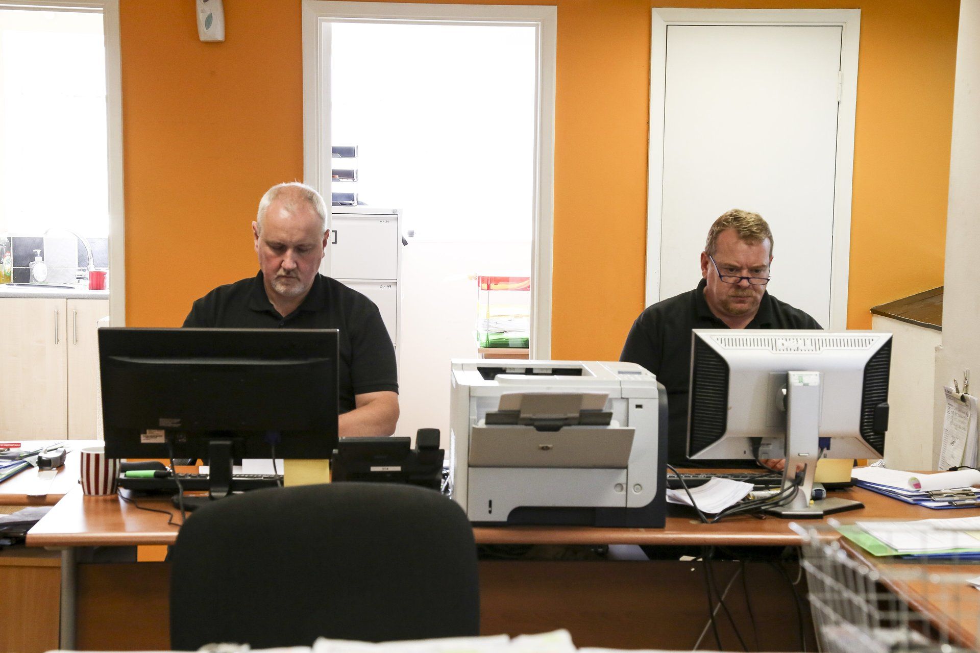 Two men are sitting at desks with computers and a printer.