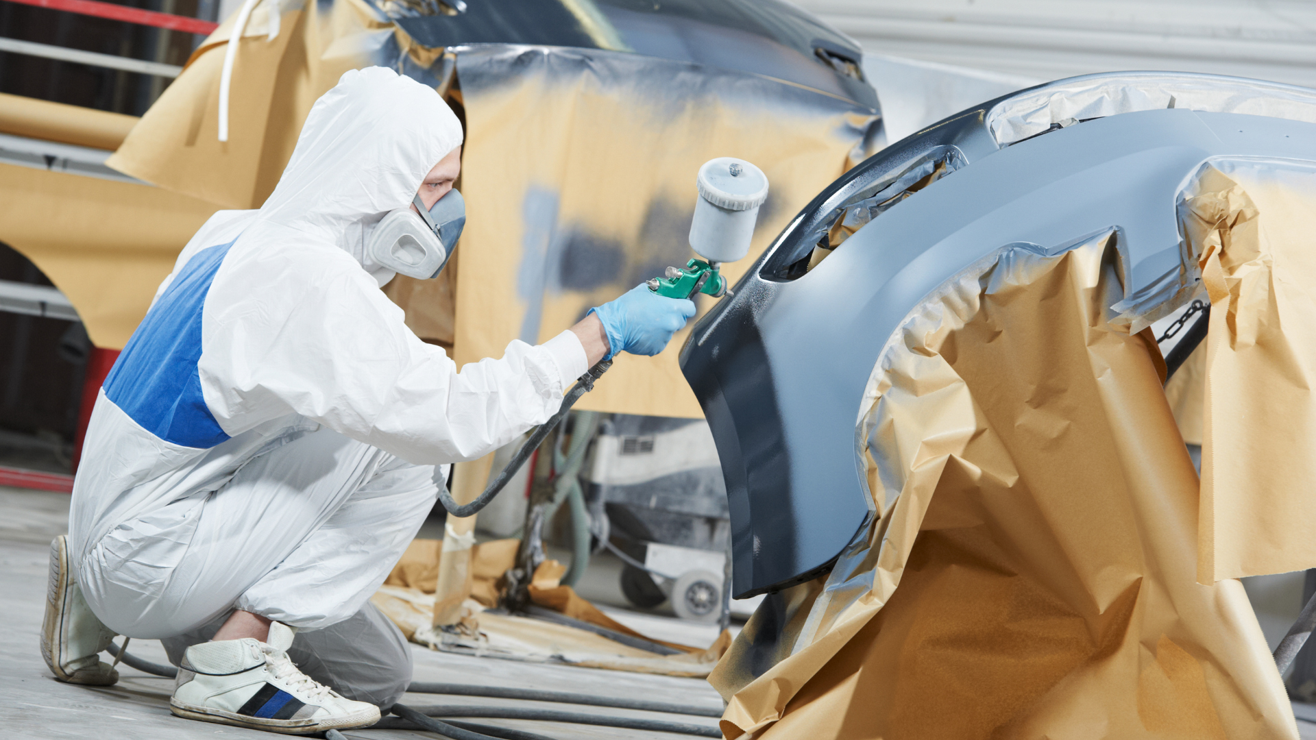 Person in protective suit spray-painting a car bumper in a workshop.
