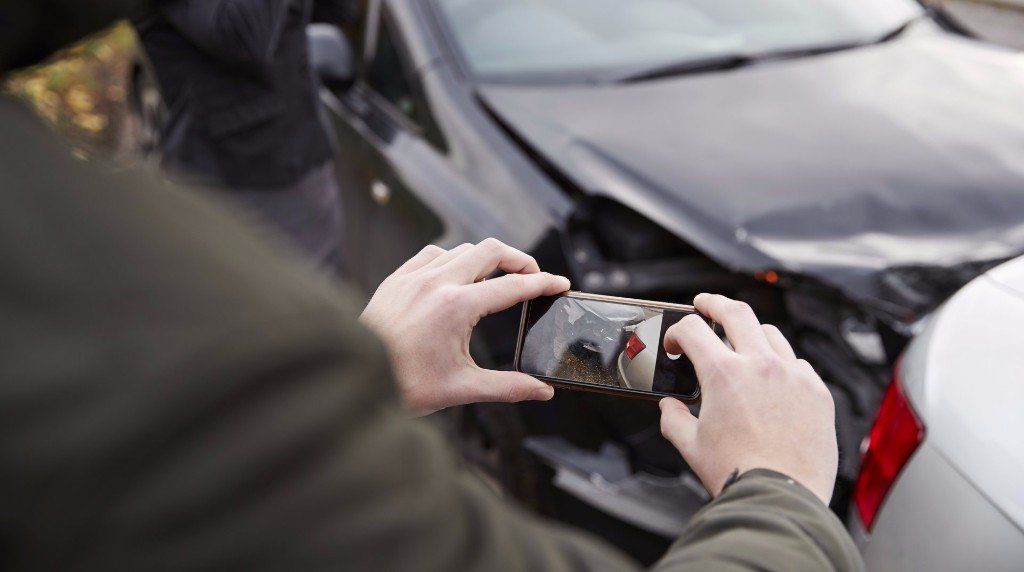 A man at a car repair workshop is taking a picture of a car accident with a smart phone.