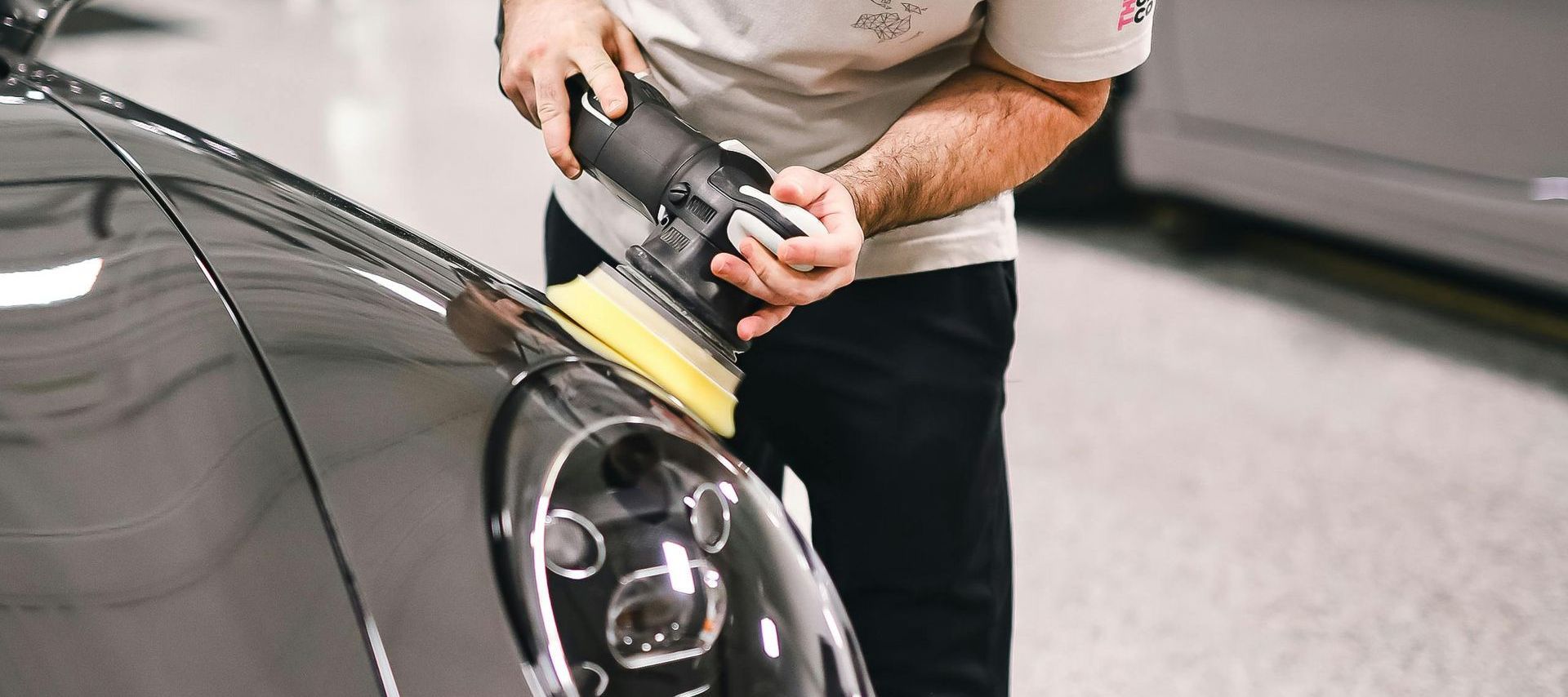 A person using a polisher on the front of a grey car in a repair shop.