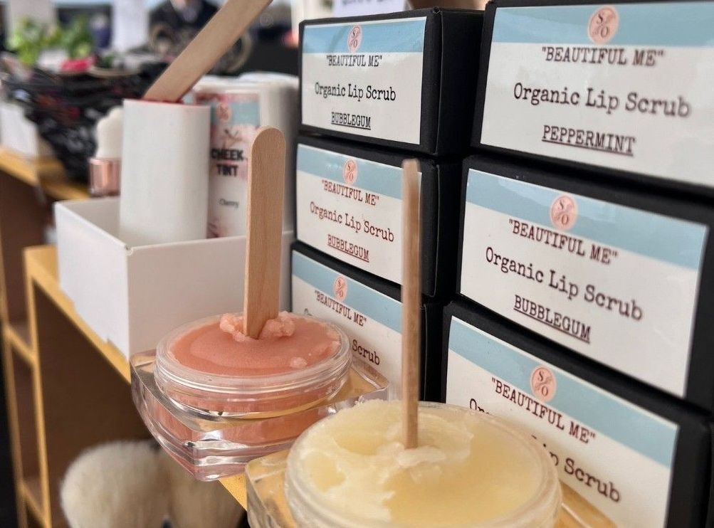 Close-up of lip scrubs in glass jars with wooden sticks, in front of boxes labeled